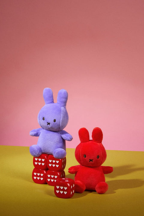 Two plush toys, Lucky Miffy, one blue and one red, on a yellow surface with a pink background.